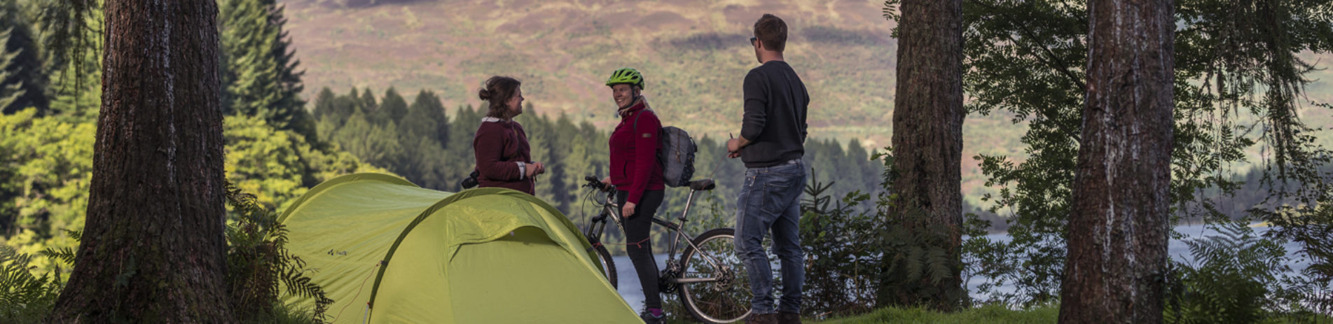 young-couple-pitching-large-green-tent-chatting-with-cyclist-on-three-lochs-forest-drive-landscape-beyond-is-of-forests-and-loch-in-the-sunshine