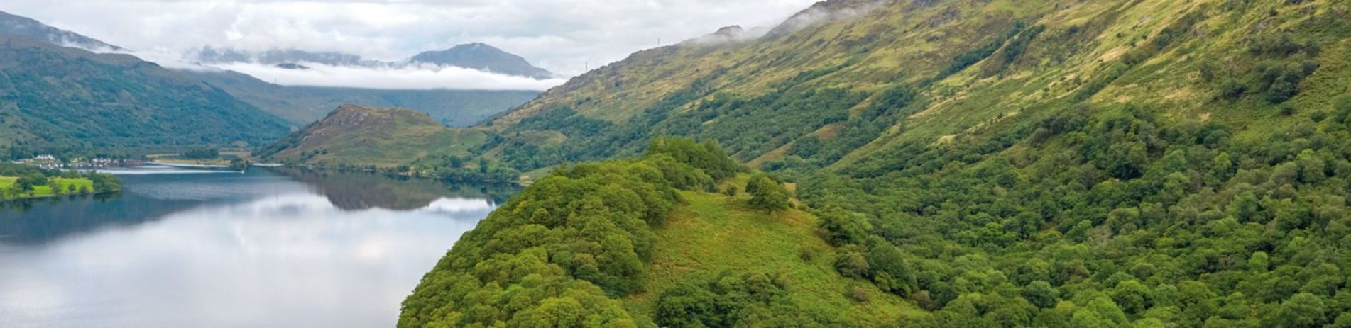 wooded-loch-lomond-shore-on-northern-end-with-mountains-peeking-in-the-distance-above-wisps-of-cloud