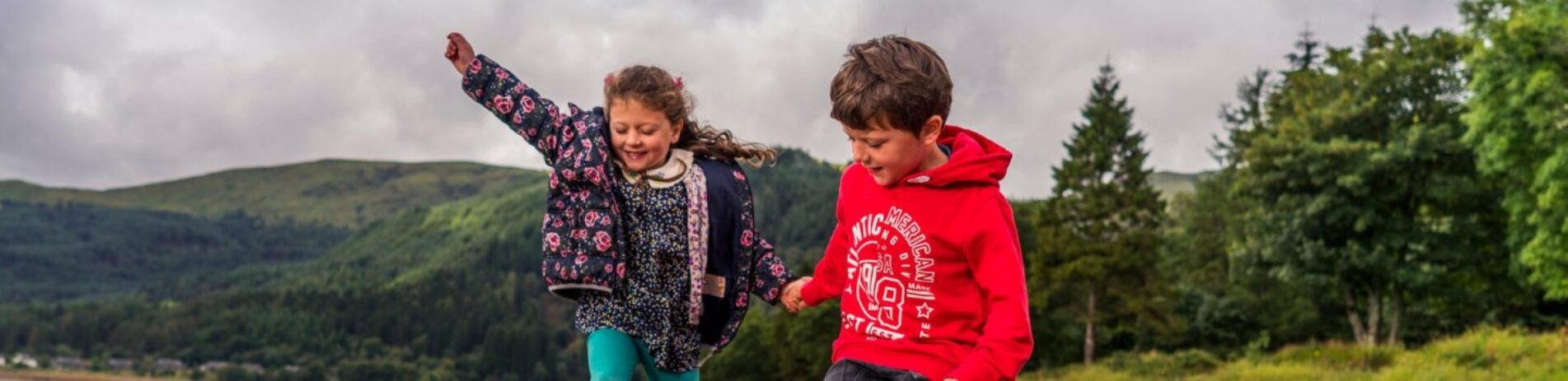 two-young-children-girl-and-boy-dressed-in-colourful-clothes-jumping-on-ardentinny-beach-with-lush-wooded-shore-of-loch-long-on-the-right-of-the-picture