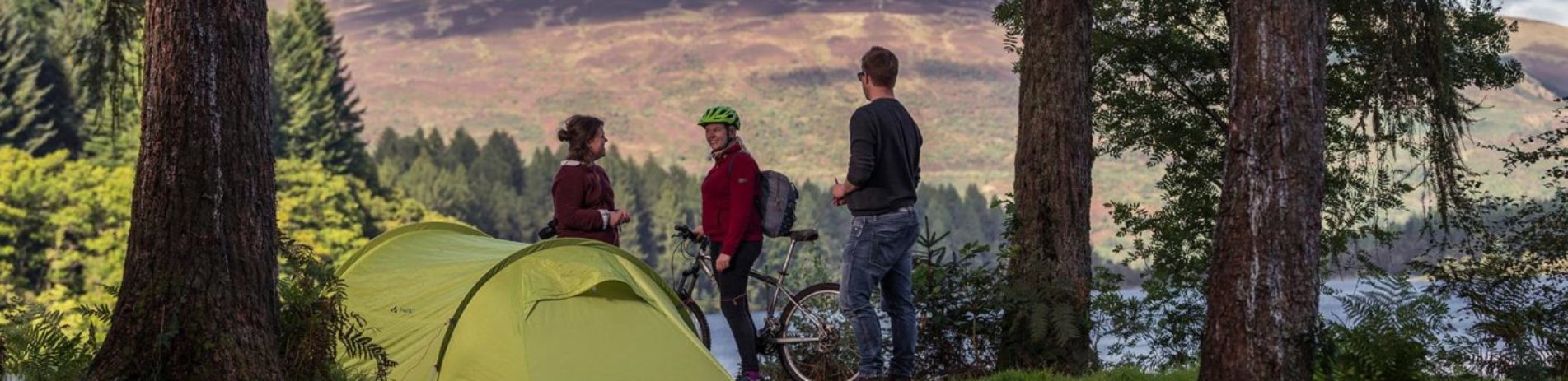 young-couple-pitching-large-green-tent-chatting-with-cyclist-on-three-lochs-forest-drive-landscape-beyond-is-of-forests-and-loch-in-the-sunshine