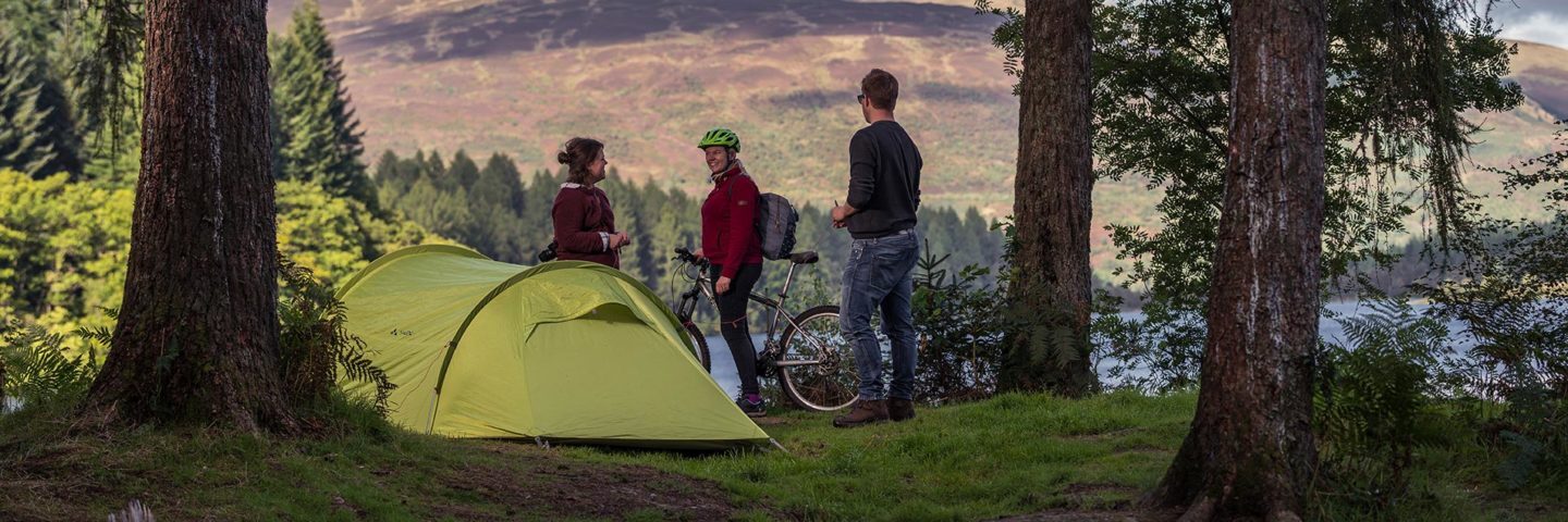 young-couple-pitching-large-green-tent-chatting-with-cyclist-on-three-lochs-forest-drive-landscape-beyond-is-of-forests-and-loch-in-the-sunshine