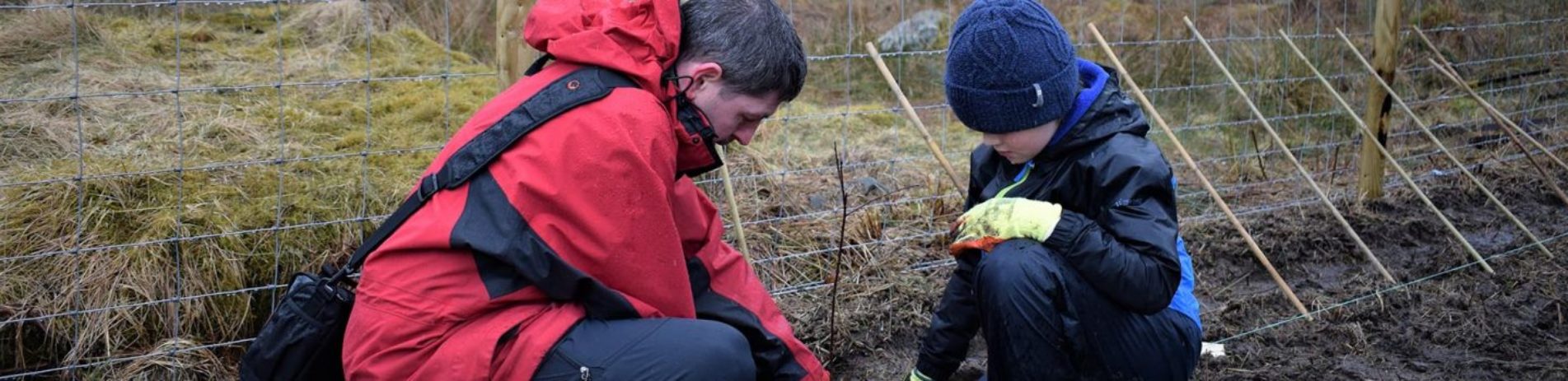 man-with-dark-hair-wearing-red-jacket-helps-young-boy-wearing-blue-hat-to-plant-a-tree