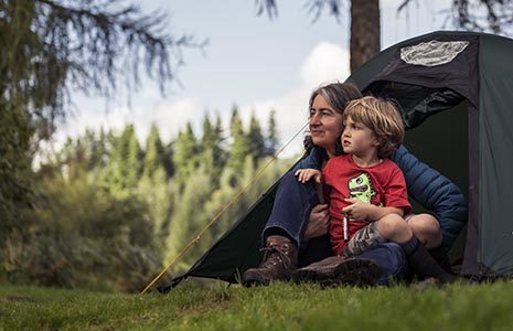 woman-and-child-at-tent-entrance-sitting-down-and-looking-in-the-distance-at-edge-of-forest-with-coniferous-trees-in-the-background