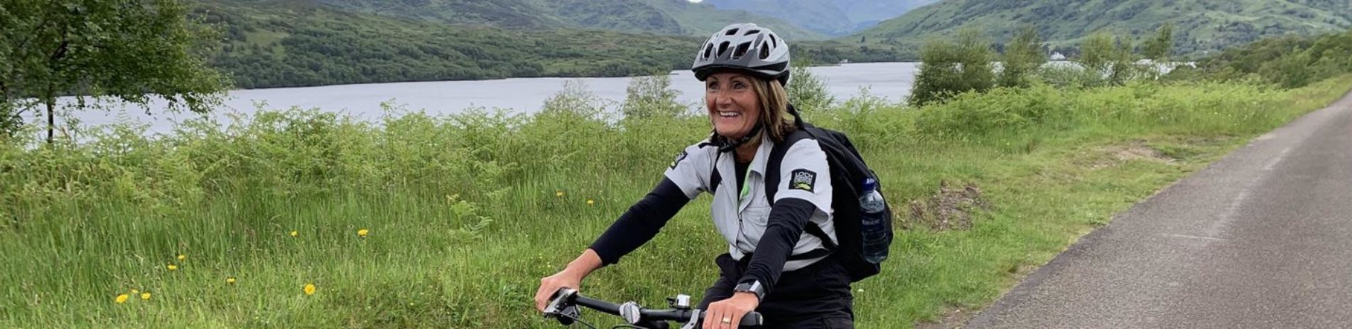 volunteer-ranger-woman-with-backpack-on-riding-smiling-with-helmet-on-with-view-of-loch-and-forested-hills-in-the-distance