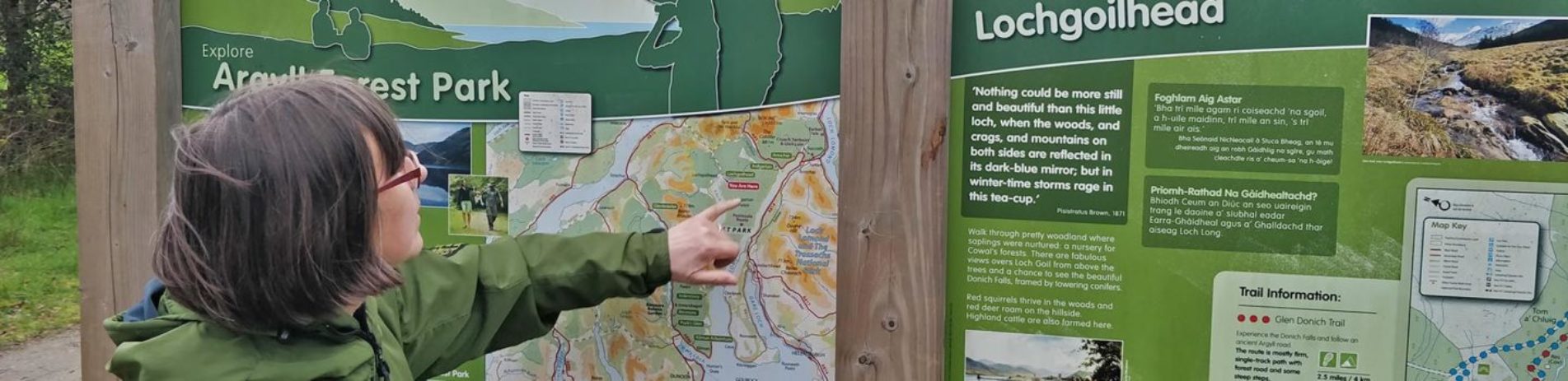 volunteer-ranger-woman-in-green-jacket-pointing-at-forestry-and-land-scotland-board-about-argyll-forest-park-and-lochgoilhead