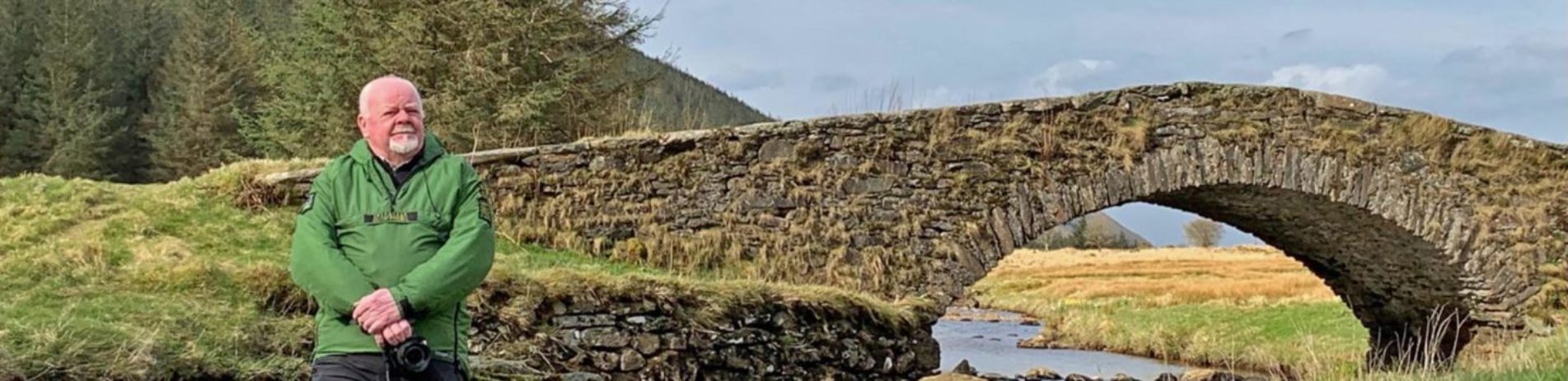 older-volunteer-ranger-male-in-green-jacket-holding-dslr-camera-posing-next-to-butterbridge-in-glen-kinglas