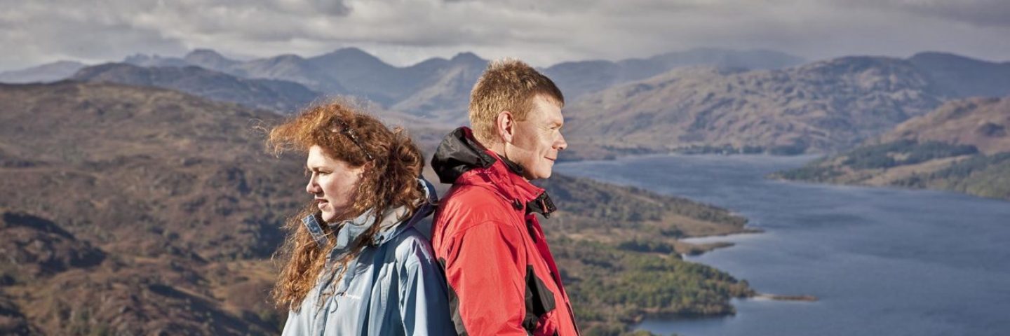 woman-and-man-in-outdoor-gear-back-to-back-admiring-views-of-trossachs-mountain-and-loch-katrine-from-summit-of-ben-aan