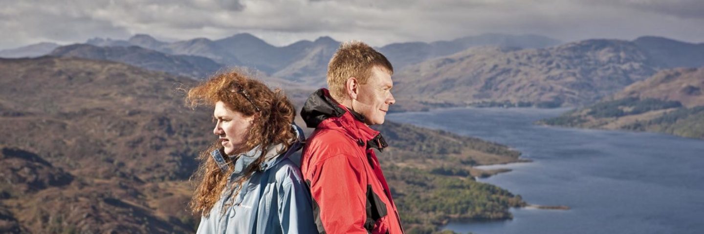 woman-and-man-in-outdoor-gear-back-to-back-admiring-views-of-trossachs-mountain-and-loch-katrine-from-summit-of-ben-aan