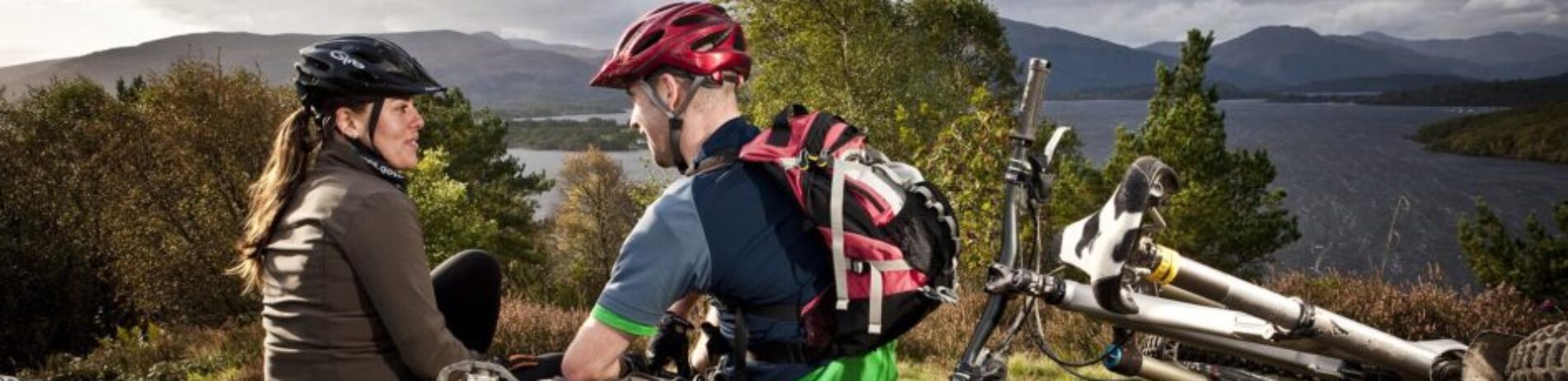 woman-and-man-in-biking-gear-with-helmets-on-next-to-their-bikes-staring-in-the-distance-over-loch-lomond