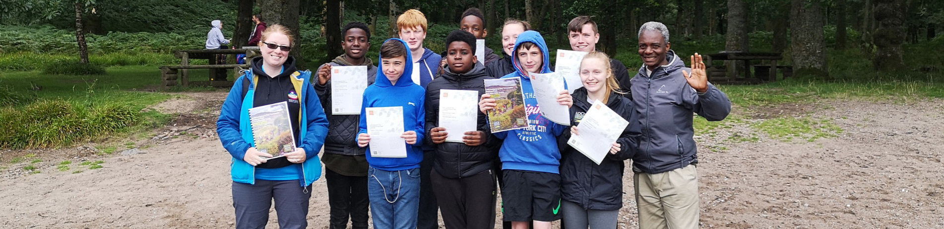 group-of-young-male-and-female-students-and-teacher-on-right-all-proudly-holding-up-certificates-standing-on-port-bawn-beach-on-inchcailloch-beach-with-forest-and-picnic-benches-behind