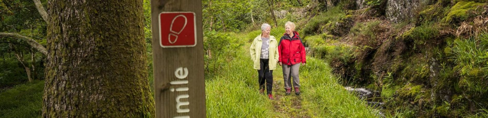 two-older-women-in-outdoor-jackets-walking-on-forest-path-in-ardentinny-distance-wooden-post-in-the-foreground