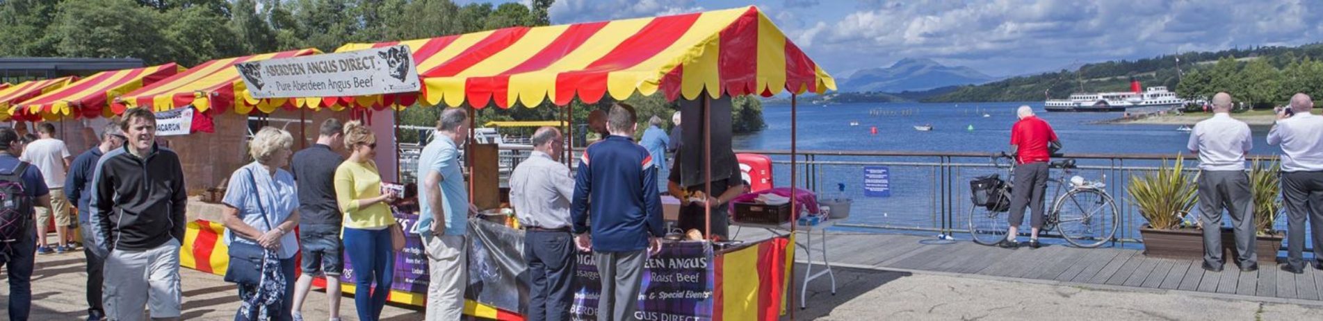 people-looking-at-food-at-red-and-yellow-stalls-at-the-edge-of-loch-lomond-in-balloch-at-food-market