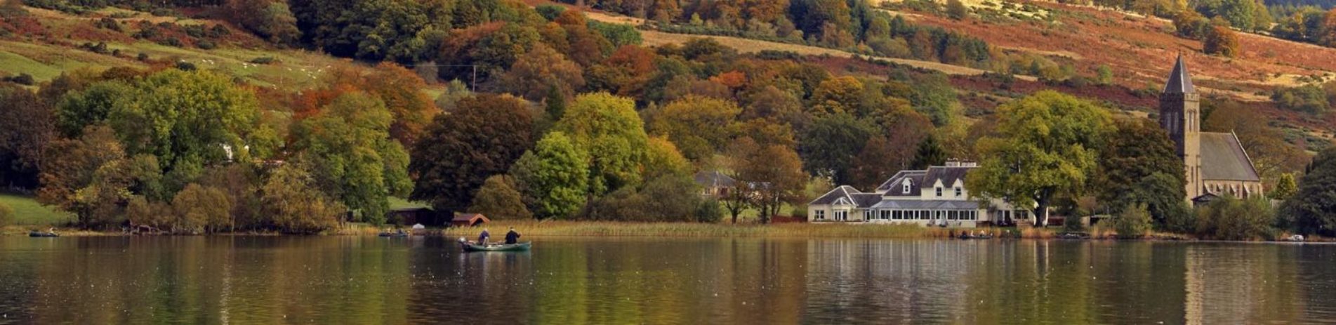 boat-on-lake-of-menteith-with-house-and-church-in-the-distance-and-autumn-colour-trees-reflected-on-the-water