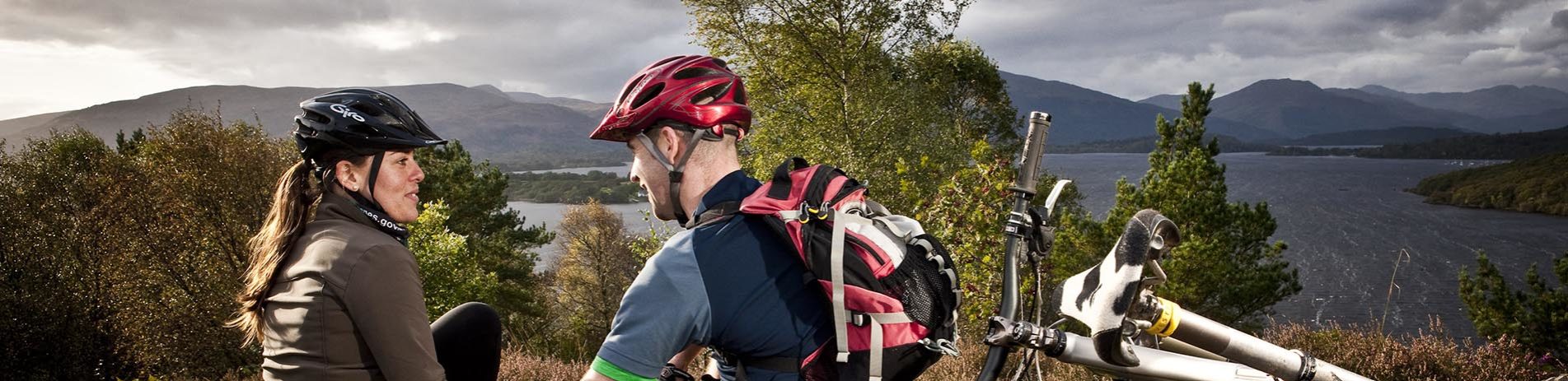 couple-wearing-helmets-with-bikes-chatting-with-loch-lomond-and-hills-in-the-distance
