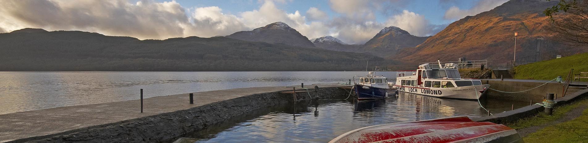 inversnaid-pier-with-two-moored-boats-and-upturned-red-boat-and-arrochar-alps-summits-in-the-distance-over-loch-lomond