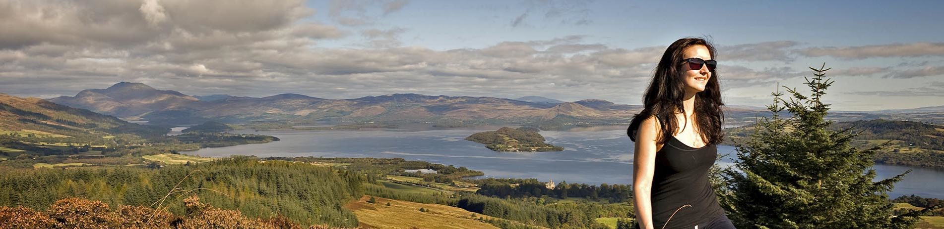 woman-walking-with-view-of-loch-lomond-ben-lomond-and-islands-in-background-and-blue-skies-partly-covered-by-clouds