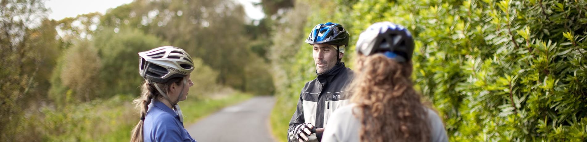 three-cyclists-wearing-bike-helmets-chatting-on-bike-path-with-trees-on-the-left-and-rhododendron-bushes-on-the-right