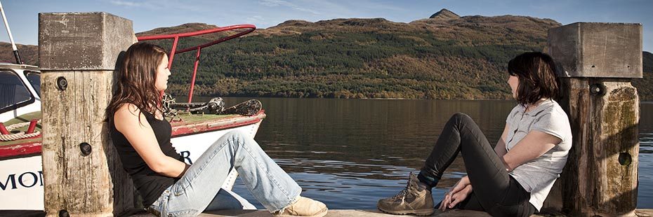 two-young-women-sitting-relaxed-on-edge-of-pier-looking-over-loch-lomond-water-and-ben-lomond-towring-abover-hills-and-forests