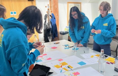A group of young people in blue hoodies stand around a table with sheets of paper and post-its.