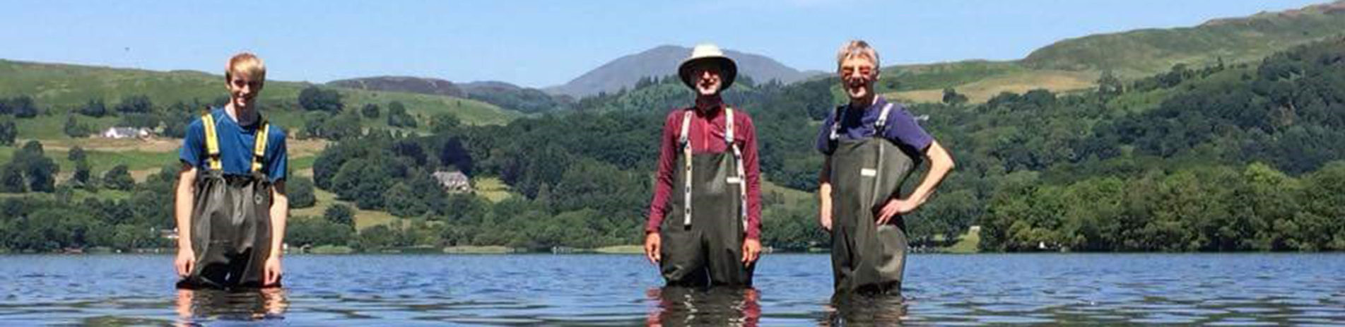 three-men-wearing-waders-in-a-loch-with-mountains-behind-on-a-sunny-day