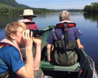 young-man-with-auburn-hair-looking-throuh-binoculars-on-a-boat-with-two-older-men-at-the-helm-backs-turned-to-camera