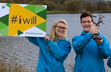 young-blonde-girl-holding-up-sign-saying-#iwill-next-to-young-brunette-boy-holding-up-medal-both-wearing-blue-youth-committee-hoodies