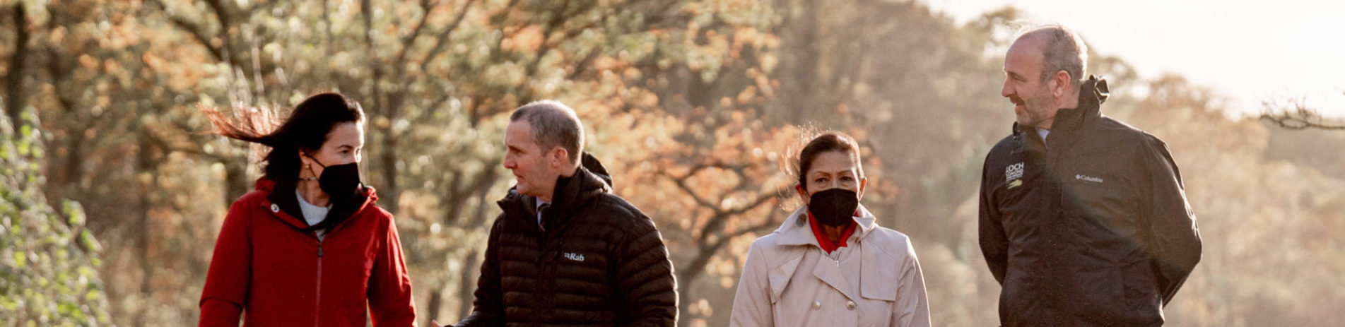 4 people walk along a pebbly beach with autumn leaves at their feet and a windswept Loch Lomond in the background