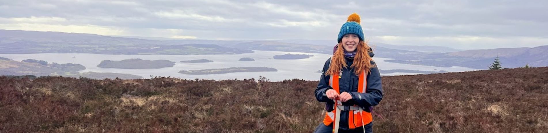 National Park staff member standing in area of peatland with Loch Lomond and islands visible in the background.
